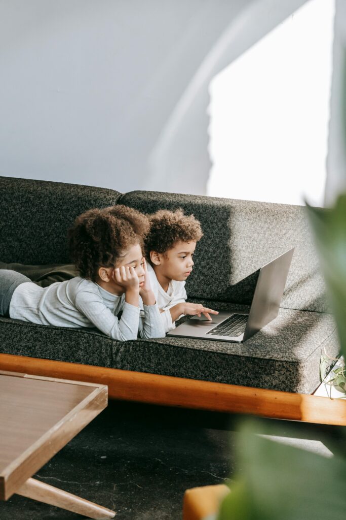 Two children lying on a sofa, focused on a laptop, enjoying tech time indoors.
